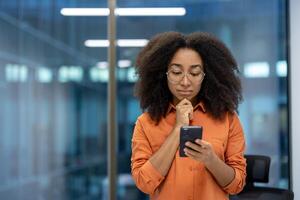 Serious thinking woman standing with phone in hands near window inside office. Businesswoman reading online news and browsing internet pages at workplace, using app. photo