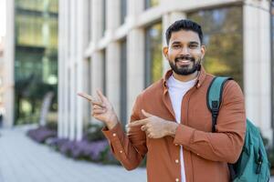 A smiling young man gestures with his hands, pointing towards an area of interest. He is wearing a backpack in front of a modern building. photo