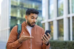 A young man outside reacts with surprise while looking at his smartphone. He gestures with his hand, seemingly in shock at the news displayed on the screen. photo