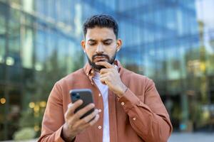 A man considers his phone in front of a modern building, contemplating the content on the screen. Thinking about communication. photo