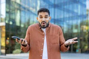 A surprised man holds a phone, reacting with a shocked expression while standing outside a modern office building. photo