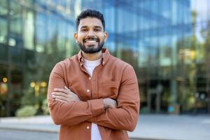 A smiling man stands confidently with arms crossed, posing in front of a modern glass building. The outdoor setting captures a professional and approachable vibe. photo