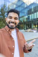 A man smiling and gesturing with his hand during a call with a modern building in the background. He is friendly photo