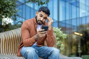 A concerned man looks at his phone while sitting outside on a bench. The scene shows a modern building in the background. photo