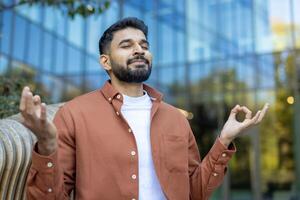 A serene man meditates with hands in a yoga mudra pose. He sits outdoors in front of a building, finding peace and tranquility. photo