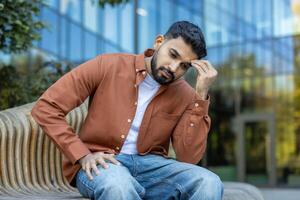 A contemplative man sits on a bench outside, deep in thought with his hand to his forehead. The modern architecture adds a sharp contrast to the scene's nature. photo
