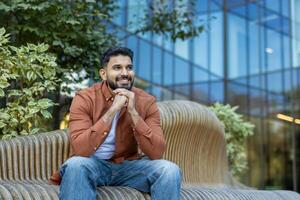 A cheerful man sits on a modern bench in front of a building, smiling and looking to the side. photo