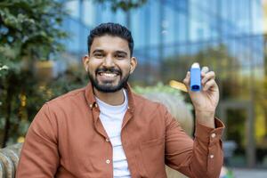 A smiling man outdoors holds up an inhaler, indicating asthma or respiratory health. The background shows a blurred building and greenery. photo