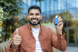 A cheerful man with an inhaler shows a thumbs-up outside. He appears to be happy and healthy, standing in front of a modern building. photo
