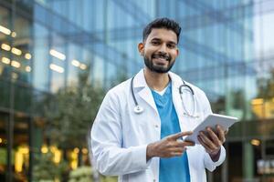 An Indian doctor smiles while using a tablet in front of a modern glass building, a scene capturing modern medicine and technology. photo