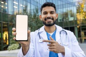 A friendly doctor holds a smartphone with a blank screen, pointing to it while smiling outside a modern building. photo