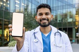 A smiling doctor holds up a phone with a blank screen, ready for your content. The medical professional stands in front of a modern building. photo
