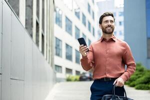A well-dressed man walks outside, holding a phone in one hand and a briefcase in the other, smiling. photo