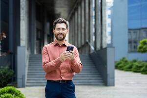 A smiling businessman holds a phone in front of a modern building, possibly checking his messages or email. The man is looking directly at the camera. photo