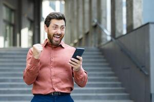 An excited man, holding his phone and cheering, stands in front of a building. The man appears happy about something he saw on his phone. photo
