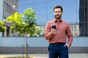 A smiling man checks his phone while standing outside near a building with reflective windows, holding a smartphone. photo