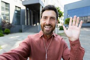 A man with a beard smiles while waving at the camera outside near a modern building, he wears earbuds. photo