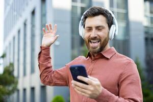 A man wearing headphones and smiling while holding a smartphone is waving in an outdoor setting. The image conveys happiness and connection. photo