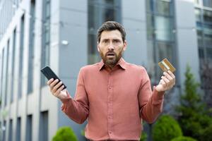 A man with a worried expression holds a credit card and a smartphone, seemingly facing a financial issue outdoors. photo