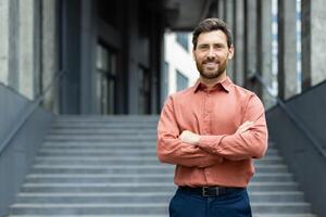 A confident man stands with arms crossed outside an urban setting, smiling directly at the camera with a cheerful disposition. photo