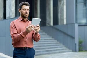 A professional man in a button-up shirt looks at a tablet in front of a modern building. He could be checking emails or reading news. photo