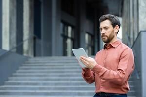 A man wearing a red shirt is focused on a tablet while standing in front of building steps. photo