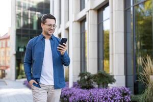 A man in glasses using his phone while walking outside, enjoying a sunny day. The urban setting includes a modern building and beautiful flowers. photo