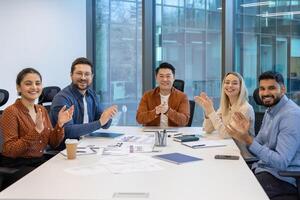 A diverse group of professionals celebrates a success during a meeting, clapping with smiles around a table. photo