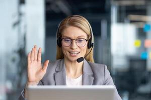 A smiling woman with eyeglasses waves during a call, working in an office environment, greeting someone with a friendly gesture. photo