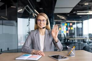A smiling businesswoman with a headset waves during a call. She is sitting at a desk with a tablet and an open notebook. photo