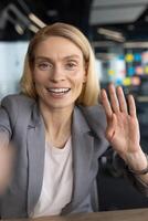 Smiling woman in a grey suit waves hello during a conference call from her office. Focus on positivity and professional connection. photo