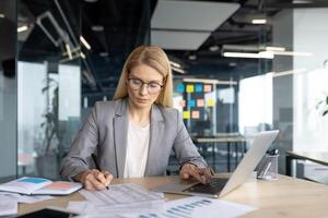 A businesswoman in a modern office setting is focused while working on a laptop and reviewing documents at her desk, analyzing data. photo