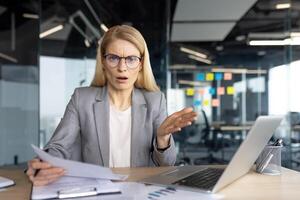 An office worker looks shocked and displeased with the documents she is reviewing while working at her desk with a laptop. photo