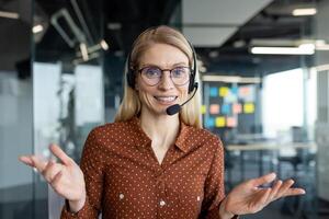 A friendly woman with glasses and a headset smiles while in an office, ready to assist. She gestures with her hands, showing openness and approachability. photo