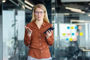 A frustrated woman in glasses holds a phone, gesturing with confusion in an office setting with glass walls and colorful sticky notes. photo