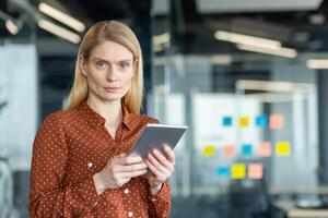 A poised businesswoman in a modern office setting, holding a digital tablet, looking directly at the camera with a composed expression. photo