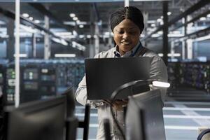 Smiling engineer using laptop in data center to oversee hardware delivering computing power for AI training models. Happy worker supervising rackmount servers storing complex machine learning datasets photo
