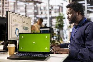 Black general manager studies corporate statistics next to chroma key, working at the office to ensure business success and growth. Male analyst working with numbers, management. photo