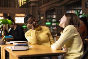 Cute teens friends gossiping together and laughing in the library, taking a break from school work and enjoying team project with each other. Young girls enjoying a cheerful conversation. photo