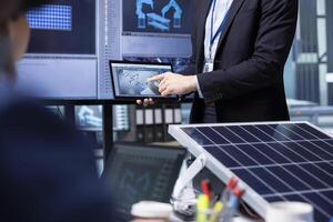 Investors examine solar panel surface using tablet, discussing design efficiency in factory office. Businessmen holding device meeting to chat about technical improvements to solar cells, closeup photo