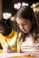 Diverse schoolgirls writing essays in a library in order to finish their homework assignments, descriptions to showcase creativity and imagination skills for the literature class. Academic growth. photo