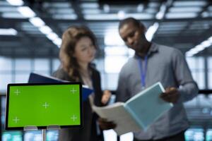 Focus on green screen tablet in data center with coworkers in blurry background employing deep learning models. Close up of mockup device used by server hub IT specialists doing brainstorming photo