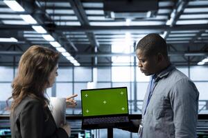 Employees in data center automating routine maintenance tasks with green screen laptop, executing diagnostic scripts. Server room workers using mockup notebook, adjusting power settings photo