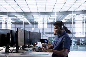 Engineer in data center listening music while using PC to check for server performance issues arising due to high CPU usage. Man wearing headphones in server farm fixing insufficient memory errors photo