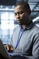 Computer scientist in data center using laptop to develop disaster recovery plans. African american man in server hub testing failover scenarios using notebook, protecting data during disruptions photo