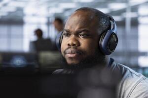 African american admin wearing headphones implementing cybersecurity measures to protect data center. Server hub worker listening music, analyzing metrics to enhance operational effectiveness photo
