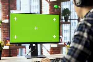 Worker listening music, analyzing data on green screen PC at desk, compiling analytical reports. Close up of employee wearing headphones using mockup computer, finishing project photo