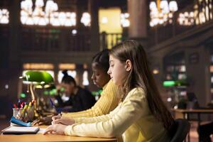 Two young students preparing for a lecture and writing ideas in notebooks at a study desk in a library, creating a reflective essay as homework. Girls focused on their school work. photo