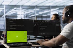 Isolated screen laptop next to man coding in server farm while listening music in headphones. Worker safeguarding hardware against unauthorized access with chroma key notebook, hearing songs photo