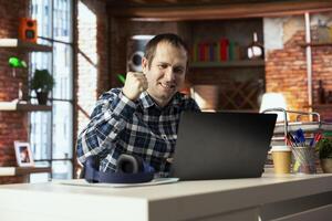 Happy worker at desk facing laptop screen, shaking fist to celebrate completion of task. Delighted teleworker in home office doing positive hand gesturing, looking at notebook screen photo